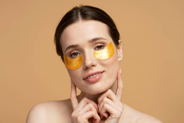Portrait of attractive smiling caucasian woman using golden under eye patches looking at camera, isolated on beige background. Skin care, morning routine, natural beauty concept - Foto, Imagen
