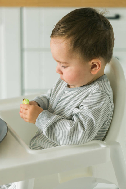Baby jongen zitten in baby stoel eten wortel op keuken achtergrond - babyvoeding - Foto, afbeelding