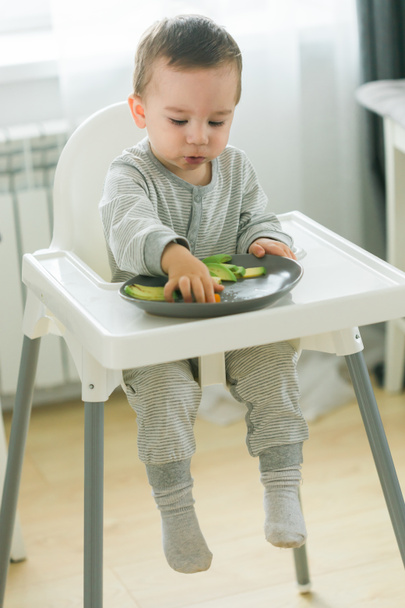 Baby boy sitting in baby chair eating carrot on kitchen background - baby feeding - Foto, Imagen