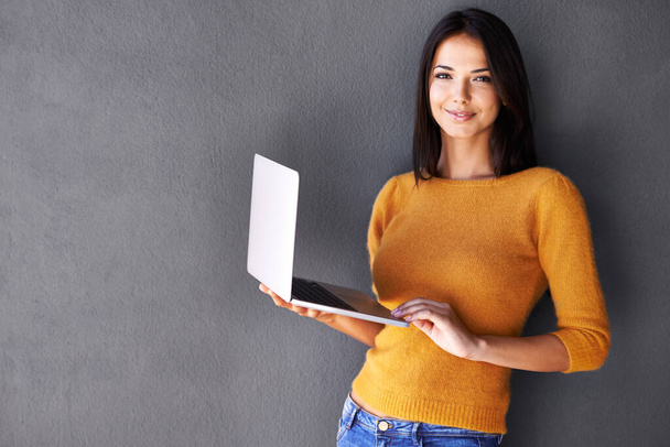 She knows how to network with people. Portrait of an attractive young woman holding a laptop while standing against a gray wall - Фото, изображение