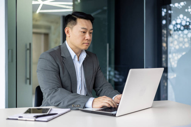 Serious and thoughtful businessman working inside office sitting at table using laptop at work, mature asian boss in shirt thinking and typing on keyboard. - Foto, Imagem