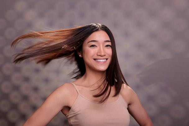 Feliz mulher asiática com cabelos brilhantes fluttering após procedimentos de rotina de cuidados com o cabelo sorrindo e olhando para a câmera. Senhora otimista confiante com retrato de penteado suave - Foto, Imagem