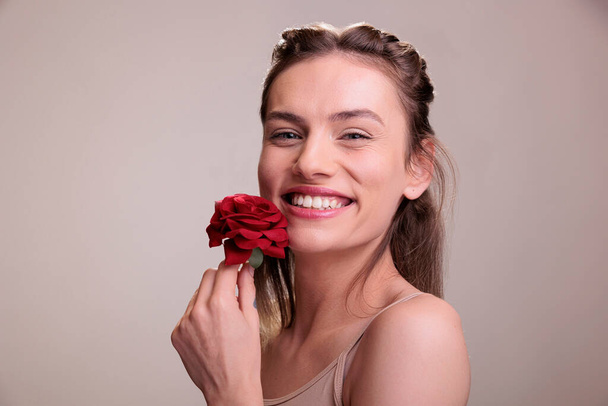 Laughing young woman posing with red rose gift portrait. Attractive cheerful caucasian blonde lady wearing nude makeup holding flower romantic present and expressing positive emotion - Photo, Image