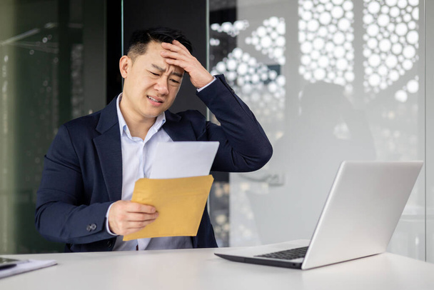 Sério e chateado asiático empresário leitura carta com más notícias, investidor homem trabalhando dentro moderno escritório edifício usando laptop. - Foto, Imagem