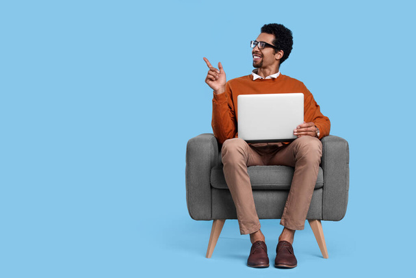 Happy man with laptop sitting in armchair and pointing at something on light blue background. Space for text - Φωτογραφία, εικόνα