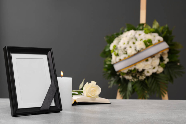 Photo frame with black ribbon, rose, burning candle on light table and wreath of flowers near grey wall indoors, space for text. Funeral attributes - Fotoğraf, Görsel