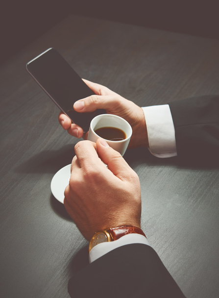 τηλέφωνο και ένα φλιτζάνι καφέ στα χέρια ενός επιχειρηματία στο σκοτάδι  - Φωτογραφία, εικόνα