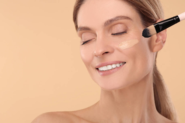 Woman applying foundation on face with brush against beige background. Space for text - Foto, Bild