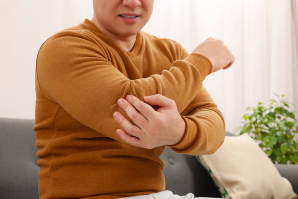 Man suffering from pain in his elbow on sofa indoors, closeup - Foto, imagen
