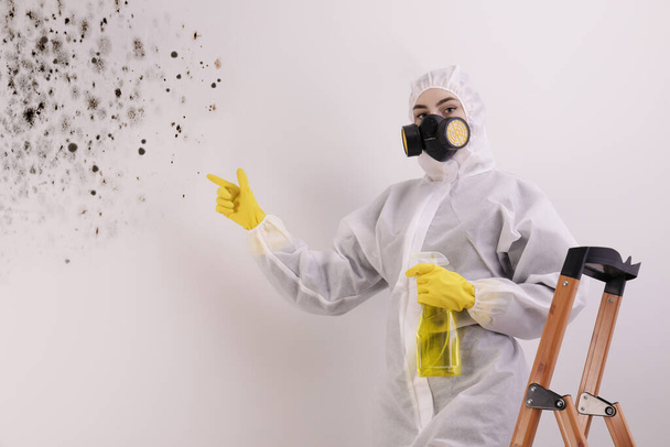 Woman in protective suit with mold remover pointing at affected wall - Foto, Bild