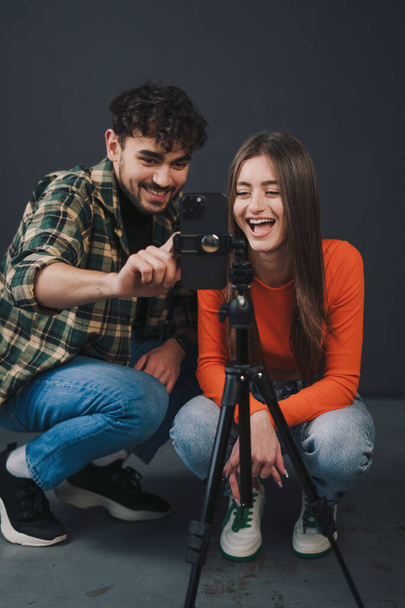 Χαρούμενος άντρας και γυναίκα bloggers που εκπέμπουν από το σπίτι, μοιράζονται τον τρόπο ζωής τους με οπαδούς, χρησιμοποιώντας smartphone σε τρίποδο απομονωμένο σε μαύρο φόντο - Φωτογραφία, εικόνα