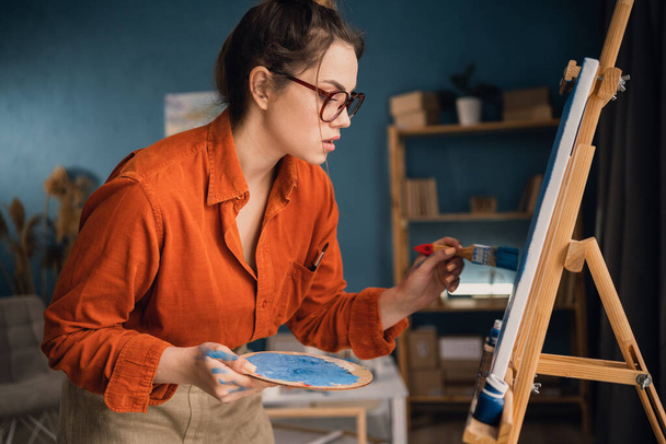 Büyük bir tuvale fırçayla resim yapan tanınmamış bir kadın sanatçı. Sanat atölyesinde çalışan genç bir ressam. Yaratıcı genç kadın yeni sanat projesi için mavi bir tablo yapıyor.. - Fotoğraf, Görsel