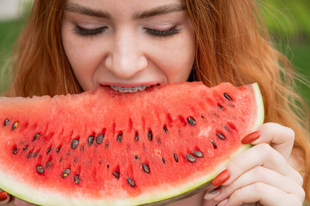 Close-up portrait of red-haired young woman with braces eating watermelon outdoors. - Fotoğraf, Görsel