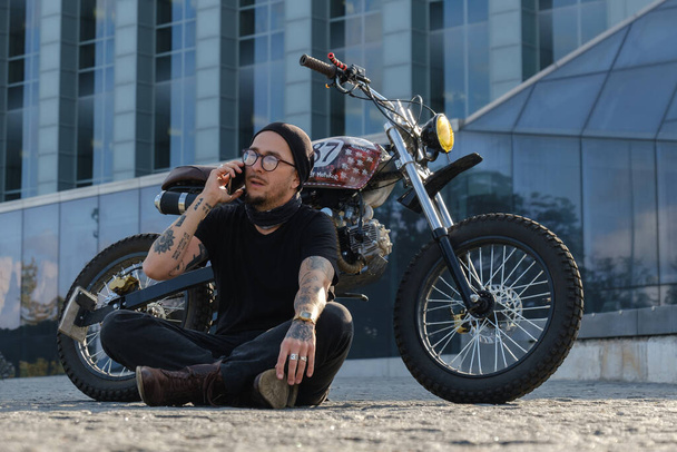 Снимок модного мотоциклиста со старинным велосипедом на фоне городского строительства. - Фото, изображение