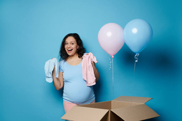Glückliche schwangere Frau mit dickem Bauch, im 2. Semester ihrer Schwangerschaft, zeigt einen blauen und rosafarbenen Neugeborenen-Body, drückt Erstaunen und Geschlecht aus, enthüllt Party, Babydusche, isoliert auf blauem Hintergrund - Foto, Bild