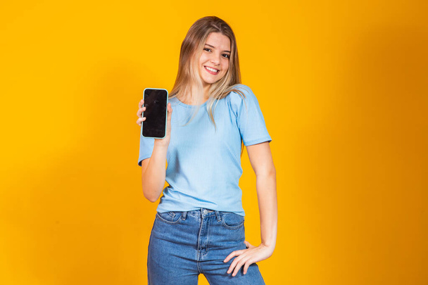 若いですブロンド女の子ショー空白のスマートフォン画面上の黄色の背景. - 写真・画像