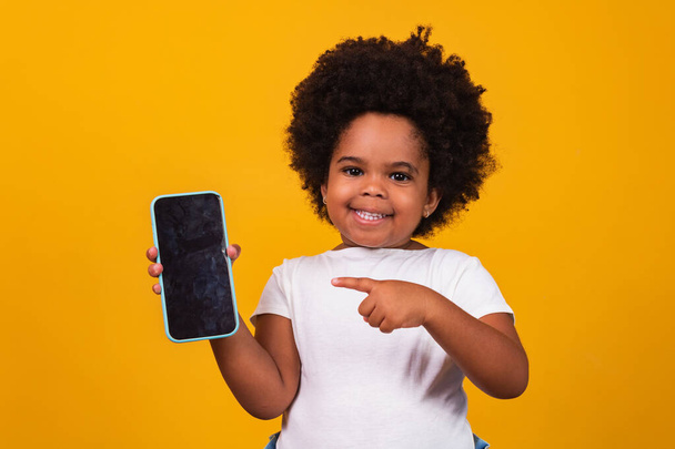 маленька дівчинка з афро, що тримає телефон і вказує ізольовано на жовтому тлі
. - Фото, зображення