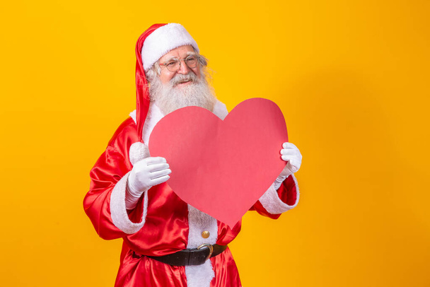 Święty Mikołaj trzyma dużą czerwoną kartę serca w żółtym tle. Romantyczny Święty Mikołaj. - Zdjęcie, obraz