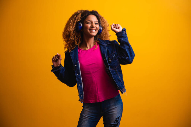 Beautiful young woman with afro hair listening to music with her headphone and dancing on a yellow background. - Fotoğraf, Görsel