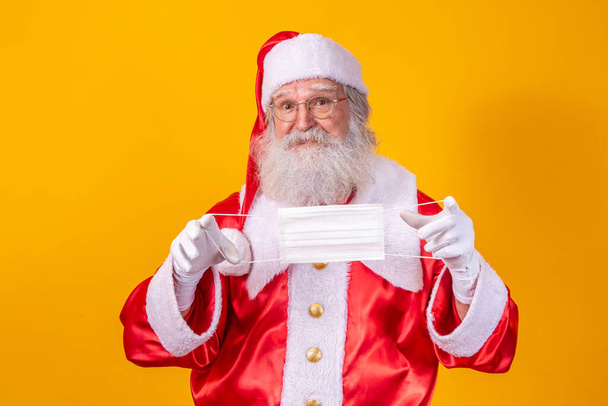 The real Santa Claus with a yellow background, holding a protective mask, glasses and hat. Christmas with social distance. Covid-19 - Photo, Image