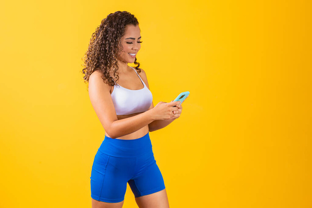 Influencer woman in fitness outfit using cell phone on yellow background - Foto, Imagen