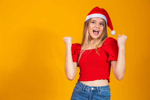 クリスマスの帽子をかぶっている若いブロンドの女の子は、腕を上げて成功したことに驚いて驚いています。勝者のコンセプト. - 写真・画像