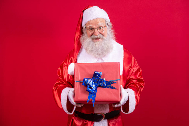 Santa Claus holding a Christmas present on red background. - Φωτογραφία, εικόνα