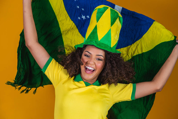 Afro girl cheering for favorite brazilian team, holding national flag in yellow background. - Foto, imagen