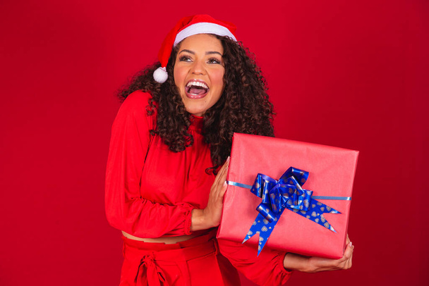 Portrait of young afro woman in santa claus hat holding a gift on red background. christmas night concept - 写真・画像