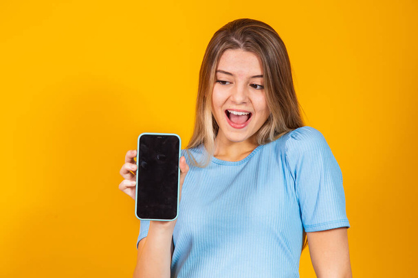 Young blonde girl showing blank smartphone screen while looking at camera with open mouth over yellow background. - 写真・画像