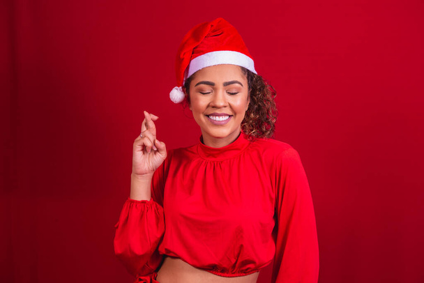 young woman in christmas outfit and santa claus hat with fingers crossed wishing luck - 写真・画像