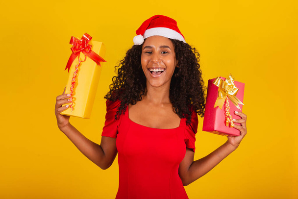 Beautiful black woman in santa claus hat holding merry christmas gift on yellow background. - Fotografie, Obrázek