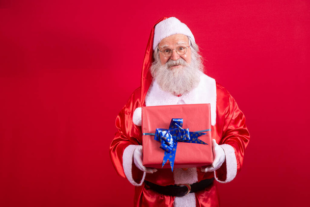 Santa Claus holding a Christmas present on red background. - Φωτογραφία, εικόνα