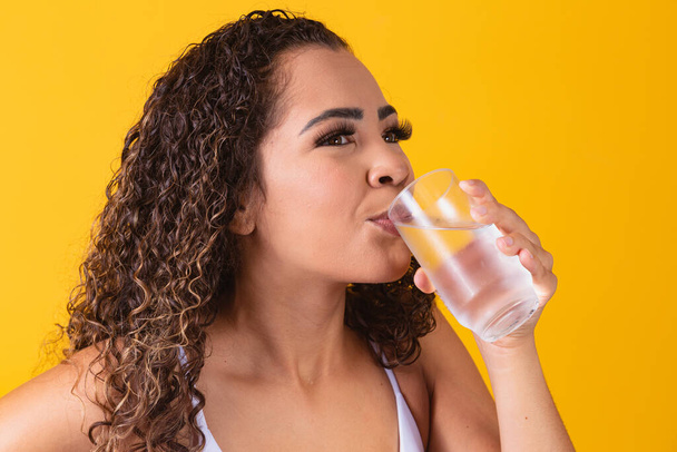 young afro woman drinking water in yellow background. Young girl with glass of water. Young woman drinking water from the glass. Hydration concept - Photo, Image
