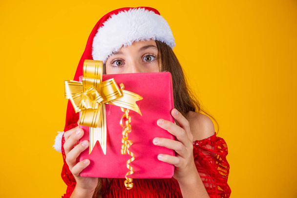Cheerful smiling funny child girl in red santa claus hat holding christmas gift in hand and hiding behind gift over yellow background. - Фото, изображение