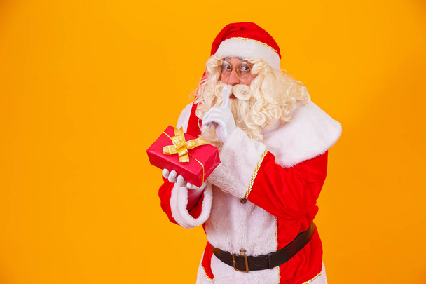 Santa Claus sobre fondo amarillo sosteniendo un regalo en las manos y haciendo una señal de silencio con la otra mano. Santa Claus sorpresa - Foto, imagen