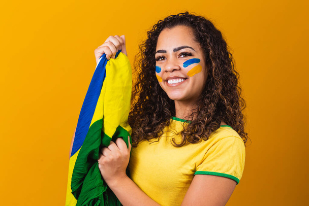 Afro girl cheering for favorite brazilian team, holding national flag in yellow background. - Φωτογραφία, εικόνα