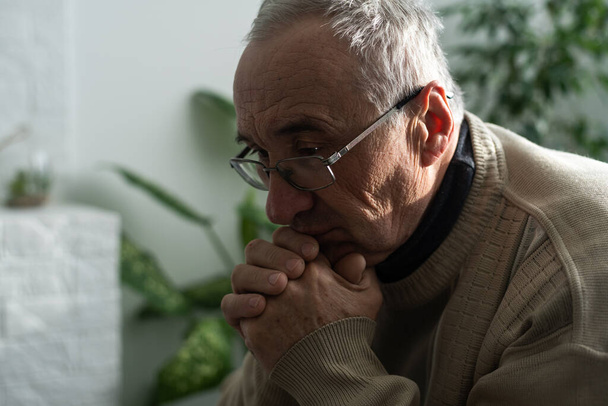 Please, Lord. Nervous worried aged Caucasian man praying on couch at home. Stressed white European senior citizen sitting on sofa, begging for forgiveness or asking God for help in difficult situation - Photo, Image