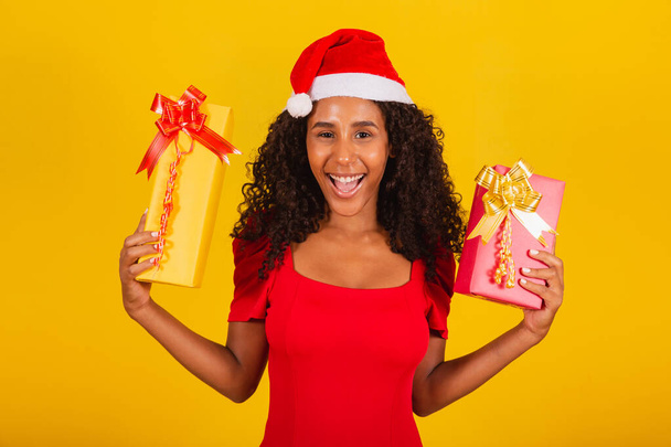 Beautiful black woman in santa claus hat holding merry christmas gift on yellow background. - Fotoğraf, Görsel