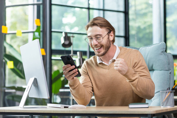 A young man works in the office at a table using a laptop. He holds the phone in his hands, looks at the screen happy and shocked, raised his glasses on his face in surprise. Got good news. - 写真・画像