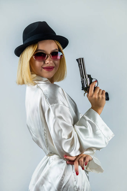 Portret seksownej eleganckiej kobiety w szlafroku, pozującej w czarnym kapeluszu i trzymającej broń. Koncepcja strzelania do studia. izolowany na równym tle - Zdjęcie, obraz