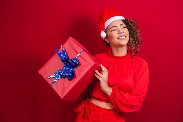 Retrato de una joven afro en sombrero de Santa Claus sosteniendo un regalo sobre fondo rojo. concepto de la noche de Navidad - Foto, Imagen