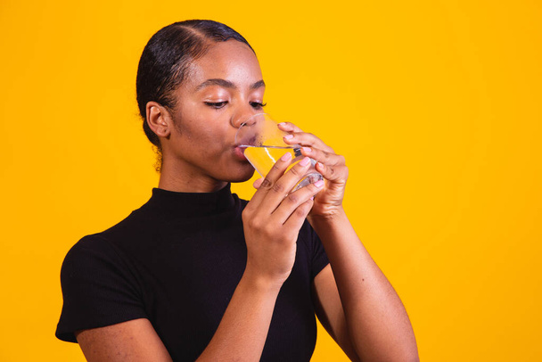 young afro woman drinking water in yellow background. Young girl with glass of water - Foto, Imagem