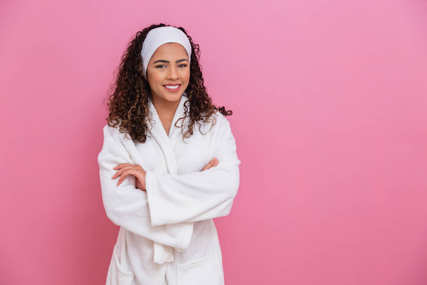 beautiful afro girl dressed in white bathrobe and sash on pink background with space for text. health and care concept. Cross arms - 写真・画像