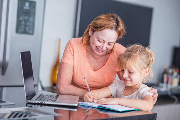 Smiling mature mother helps her daughter in preparing chores at home. Online education concept. - Foto, Bild