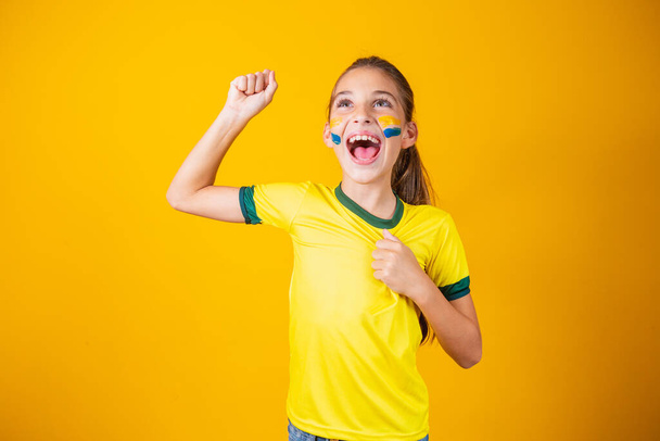 Beautiful little girl rooting for her team on yellow background. Little girl celebrating the goal and celebrating Brazil's victory - Valokuva, kuva