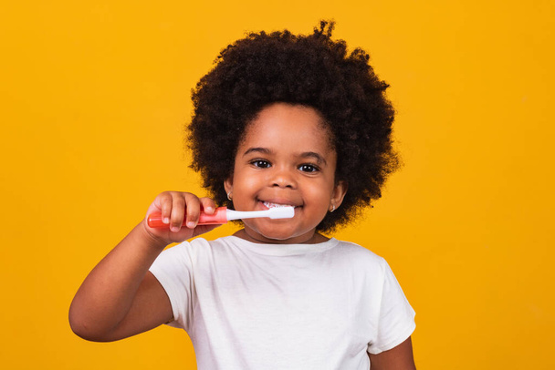 A little cute african american girl brushing her teeth, isolated over yellow background. Healthy teeth concept. - Foto, Imagem