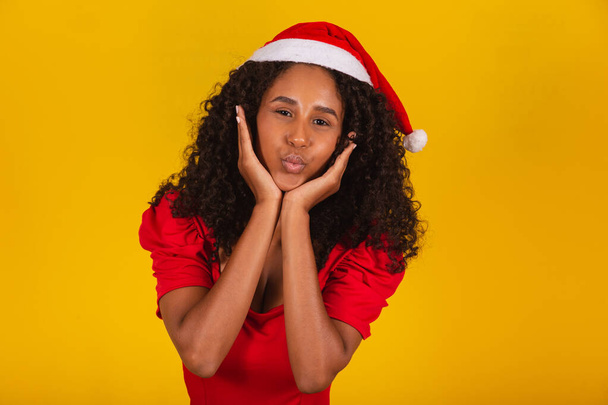 detailní záběr afro žena na sobě Santa Claus klobouk pokroucené rty trucování nebo foukání polibky na žlutém pozadí - Fotografie, Obrázek