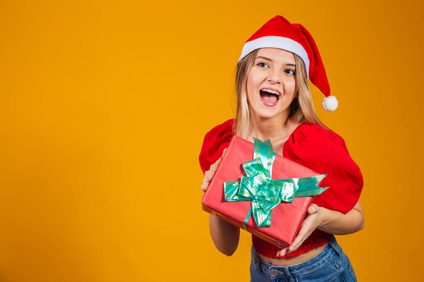 Smiling excited woman in red santa claus outfit holding christmas gift at her ear isolated on the yellow background - Φωτογραφία, εικόνα