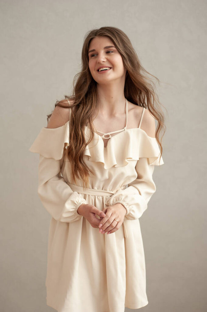 Hermosa joven mujer sonriente retrato con el pelo ondulado, vestido con un vestido beige posando en el interior  - Foto, imagen